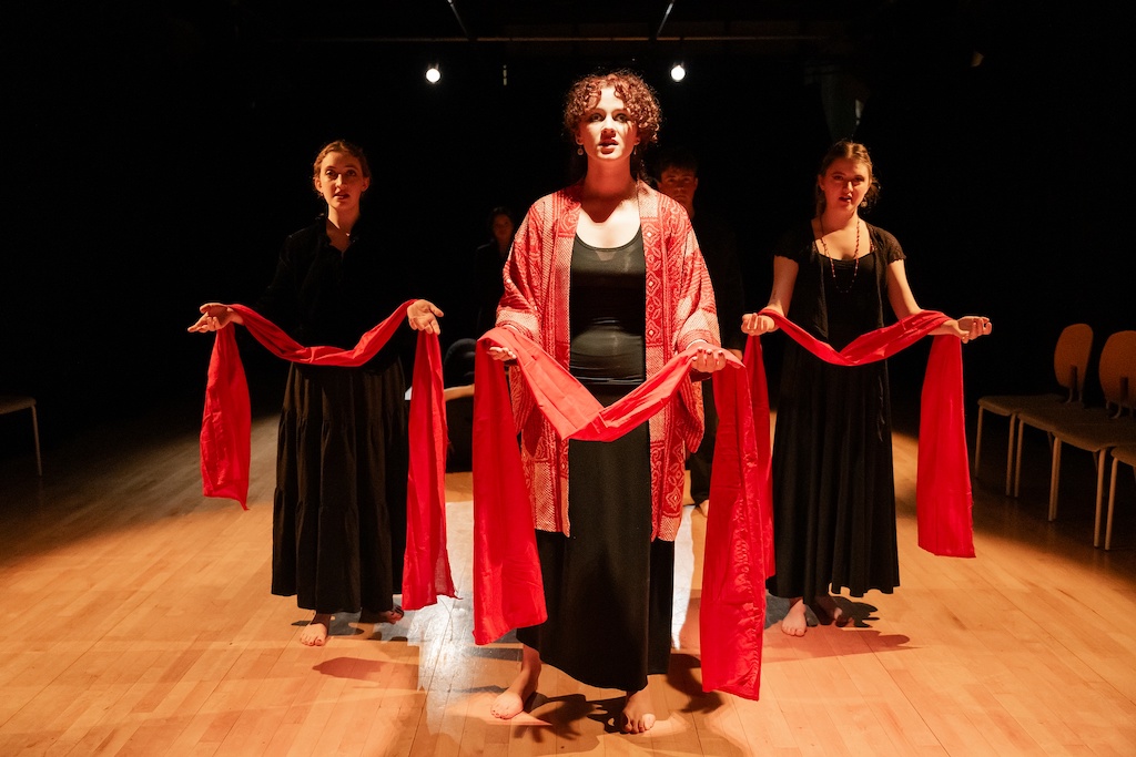 Three actors wearing long black dresses stand on stage holding bright red capes. The actor in the center is also wearing a patterned light red coat over her dress.